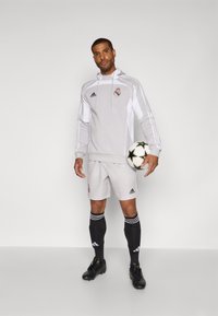 Sivi adidas dukserica s bijelim prugama, sportske kratke hlače, crne čarape i kopačke. Drži nogometnu loptu s uzorkom zvijezda. Na odjeći se nalazi logotip.
