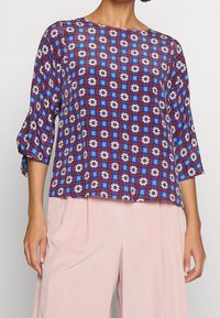 Blouse à motifs floraux avec des couleurs bleu, rouge et blanc, à coupe carrée et manches trois-quarts, associée à un pantalon rose clair.