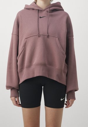 Personne portant un sweat à capuche Nike mauve oversize et un short cycliste Nike noir, debout devant un fond blanc uni.