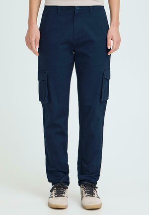 Blå cargobukser med sidelommer med klaff, båret med beige og svarte joggesko på en stående person mot en ensfarget bakgrunn.