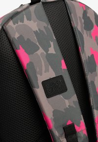 Correa de mochila en camuflaje gris y rosa con tela texturizada y respaldo de malla; presenta una etiqueta negra "SUPER DRY" en la base.