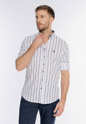 Mężczyzna poprawiający kołnierz białej koszuli z krótkim rękawem z pionowymi paskami i małymi kolorowymi kropkami, ubrany w ciemnoniebieskie spodnie, na białym tle.