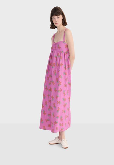 Jeune personne portant une robe maxi sans manches rose avec des motifs floraux orange et des baskets blanches, debout devant un fond clair uni.