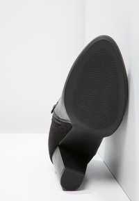 Scarpa nera con tacco alto, realizzata con una tomaia in suede texturizzato e accenti in pelle liscia. La suola è in gomma con un motivo a onde per una migliore aderenza.