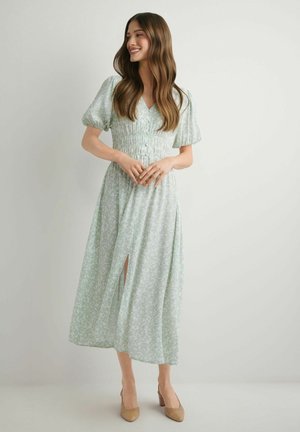 Młoda kobieta z długimi falującymi włosami, ubrana w jasnoróżową sukienkę midi w kwiaty z bufkami i beżowe szpilki, uśmiechająca się i stojąca na tle gładkiej ściany.