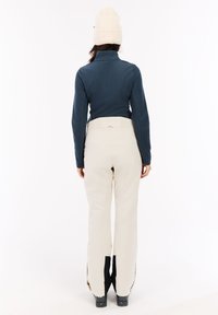 Maglione in pile blu con colletto alto, abbinato a pantaloni da neve color crema con accenti neri e zip laterali, indossato con un berretto a maglia color crema.