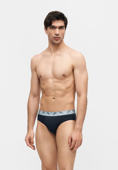 Νέος άνδρας στέκεται μπροστά σε απλό φόντο, φοράει μαύρα εσώρουχα με γκρι ζωνάκι με σχέδια, κοιτάζει μπροστά με ουδέτερη έκφραση.