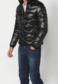 Uomo che indossa una giacca trapuntata nera lucida con tasche con zip e jeans blu, in piedi contro uno sfondo chiaro uniforme.