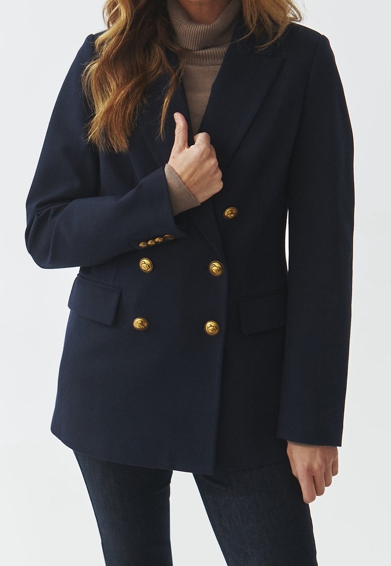 Femme portant un blazer bleu marine croisé à boutons dorés sur un pull col roulé beige et un jean foncé.