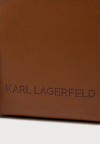 Acessório de couro castanho com a marcação perfurada "KARL LAGERFELD". Textura suave, formato retangular, design minimalista.