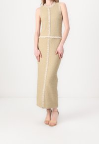 Robe beige sans manches mi-longue en tissu texturé avec bordure à franges le long des coutures, présentant une silhouette droite et un décolleté discret.