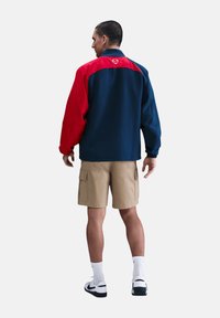 Navy und rote Windbreaker-Jacke mit hohem Kragen und einem Logo auf der Rückseite. Kombiniert mit beigen Cargo-Shorts und weißen Socken.