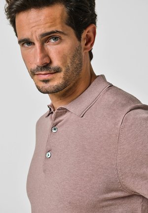 Mężczyzna z krótkimi ciemnymi włosami i brodą, ubrany w jasnoróżową koszulkę typu polo zapiętą na guziki, patrzący lekko na bok na tle jednolitego tła.