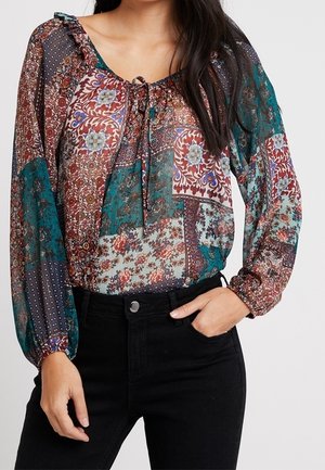 Kvinde iført en gennemsigtig, langærmet blomsterpatchwork-bluse med bindesnørred ved halsen og sorte bukser med høj talje, hånd i lommen.