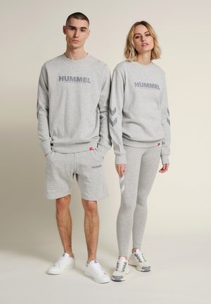 Joven hombre y mujer con sudaderas grises a juego de Hummel; hombre con pantalones cortos, mujer con leggings, ambos con zapatillas blancas.