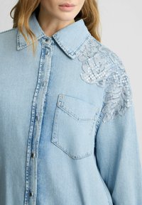 Camicia di jeans azzurro chiaro con chiusura a bottoni, colletto appuntito e ricamo floreale con paillettes sulla spalla. Taschino sul petto visibile.