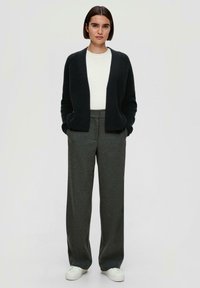 Schwarze, gerippte Strickjacke mit offenem Vorderteil, getragen über einem weißen Hemd und grauen weit geschnittenen Hosen. Das Outfit wird mit weißen Turnschuhen abgerundet.