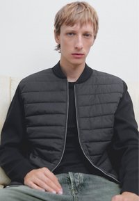 Schwarze Bomberjacke mit einem gesteppte Vorderseite, Reißverschluss und geripptem Kragen, kombiniert mit einem schwarzen Shirt und ausgewaschenen grünen Jeans.