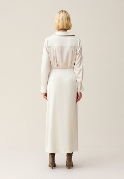 Donna in piedi di spalle, che indossa una lunga blusa crema e una gonna abbinata con stivali beige con tacco, su uno sfondo neutro.