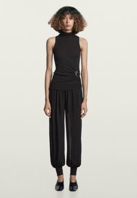 Svart ärmlös topp med hög hals och bältesdetalj i sidan; kombinerad med löst sittande, svarta koniska byxor som samlas vid anklarna.