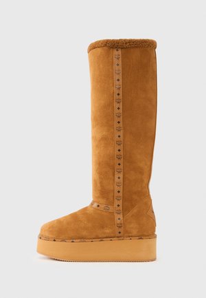 Botte hauteur genou en daim couleur camel avec un revers en peau lainée, une plateforme de 3 cm et une sangle décorative avec un logo orné de motifs en forme de diamant sur le côté.