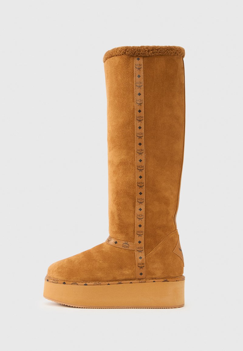 Botte hauteur genou en daim couleur camel avec un revers en peau lainée, une plateforme de 3 cm et une sangle décorative avec un logo orné de motifs en forme de diamant sur le côté.