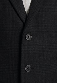 Blazer de lana negra con un sutil patrón de cuadros, solapas con muesca y dos botones oscuros en la parte delantera para el cierre.