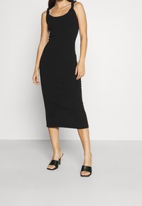 Schwarzes, geripptes Strick-Midi-Kleid mit dicken Trägern, Rundhalsausschnitt und seitlichem Schlitz. Kombiniert mit schwarzen, offenen Blockabsatzschuhen.