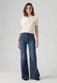 Light beige short-sleeve polo shirt paired with wide-leg dark blue jeans, showcasing a classic fit and textured fabric. Black shoes complete the look.