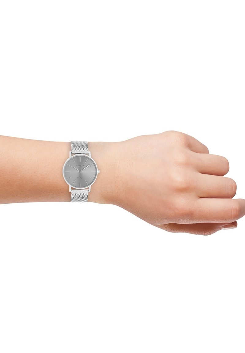 Silver mesh strap watch with a round gray dial, minimal hour markers, and slim profile, worn on a wrist, showcasing its sleek design.