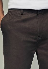 Pantalons marron foncé avec une texture lisse, une coupe ajustée, deux poches latérales et une fermeture à bouton à la taille.