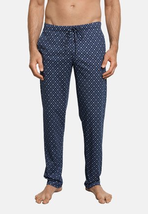 Homme portant un pantalon de pyjama bleu marine avec des motifs géométriques blancs, debout pieds nus sur un fond blanc.