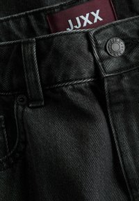 Κοντινό πλάνο στη μέση μαύρων τζιν denim με κουμπί και εμφανή κόκκινη ετικέτα με την ένδειξη JJXX μέσα στον θηλιά της ζώνης.