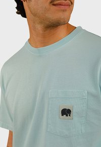 Lichtblauwe katoenen T-shirt met een ronde halslijn en een borstzak met een grijs label waarop een grafische afbeelding van een zwarte olifant staat.