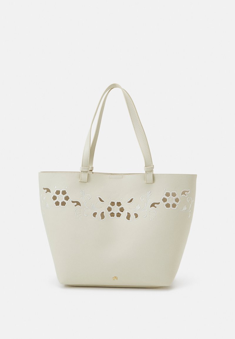 Sac fourre-tout beige avec motifs floraux découpés et broderie, avec doubles bretelles, sur un fond clair uni.