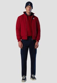 Giacca a vento rossa con colletto alto, felpa blu navy sotto, pantaloni cargo blu navy e berretto blu navy. Presenta un logo sulla giacca.
