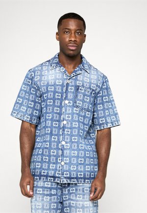 Mies, jolla on lyhythihainen sininen farkkupaita ja siihen sopivat shortsit valkoisella geometrisella kuviolla, seisoo yksinkertaista vaaleaa taustaa vasten.