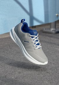 Szürke és kék sportcipő fehér fűzőkkel és talppal, aszfalt fölött lebegve, egy kék fal és egy fémpózna mellett.