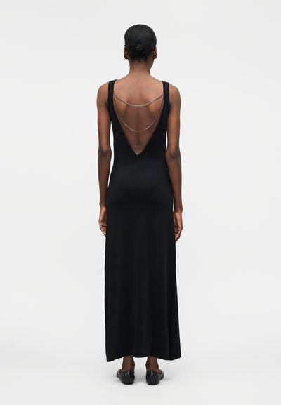 Frau mit Haaren zu einem Dutt gebunden, die ein ärmelloses schwarzes Maxikleid mit tiefem V-Rücken und filigranem silbernem Kettendetail trägt, stehend vor einem einfarbigen Hintergrund.