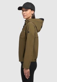 Giacca leggera verde oliva con cappuccio, polsini elasticizzati e una tasca frontale. Abbinata a un cappellino nero, il modello è in posa di profilo su uno sfondo chiaro.