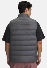 Gray padded vest with high collar and horizontal stitching. Lightweight material, features logo on the lower right, paired with a black shirt.