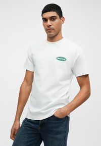 Balta medvilninė marška, kurią puošia mažas žalias logotipas kairėje krūtinės pusėje. Derinama su tamsiai mėlynomis džinsomis, kurie parodo laisvą fasoną.