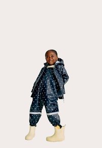 Granatowa wodoodporna kurtka i pasujące ogrodniczki w białe kropki, zestawione z żółtymi gumowcami; gładka faktura, luźny krój, refleksyjne akcenty.