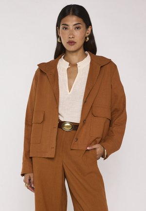 Donna con capelli lunghi e scuri che indossa una camicetta bianca, una giacca marrone, pantaloni marroni abbinati, orecchini a cerchio dorati e una cintura marrone con una grande fibbia ovale.