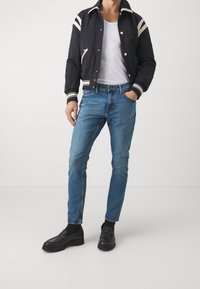 Černá bunda s bílými pruhy, bílý tričko, světle modré džíny a černé mokasíny. Bunda má zip a žebrované manžety.