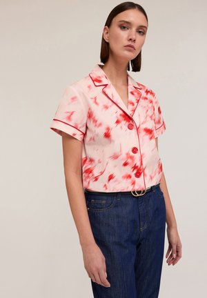 Korte mouwen, roze blouse met rood abstract patroon en rode bies, met een kraag en knoopsluiting aan de voorkant. G gedragen met blauwe jeans.