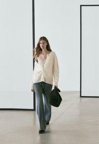 Light beige cable-knit cardigan, white lace top, dark flared jeans, and black handbag. Smooth textures, relaxed fit, minimalistic background.