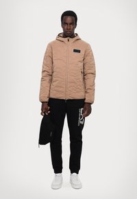 Beige quiltet jakke med hætte og lomme med sort accent; båret over sorte joggers med hvidtlogo og parret med hvide sneakers.