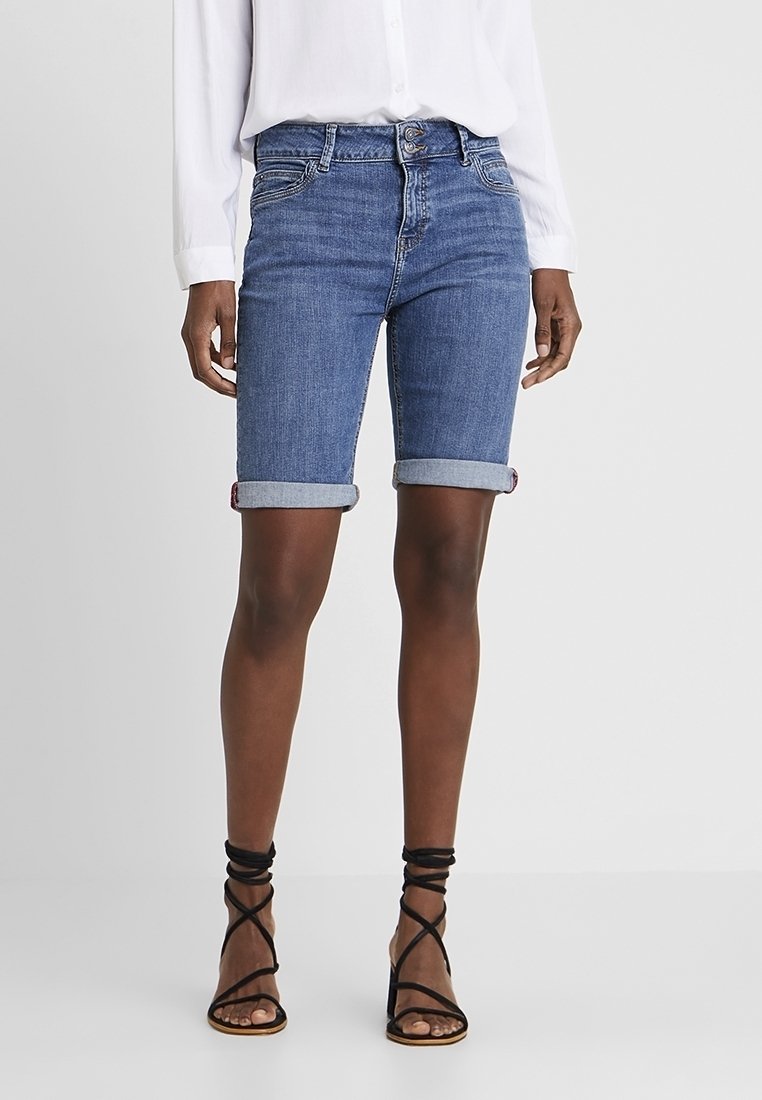 Mujer con shorts de mezclilla azul hasta la rodilla remangados, camisa blanca de manga larga y sandalias negras de tacón alto con tiras, de pie.