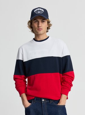 Giovane uomo con berretto blu navy con scritta bianca, felpa a blocchi di colore bianca, blu navy e rossa, e jeans blu scuro, in piedi con le mani nelle tasche.